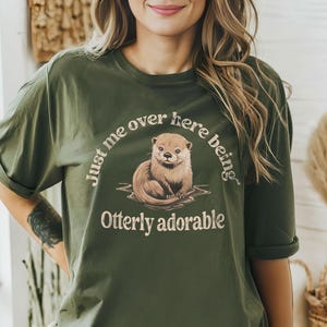 May include: A green t-shirt with a cute otter graphic and the text "Just me over here being otter-ly adorable".