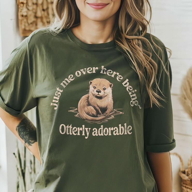 May include: A green t-shirt with a cute otter graphic and the text "Just me over here being otter-ly adorable".
