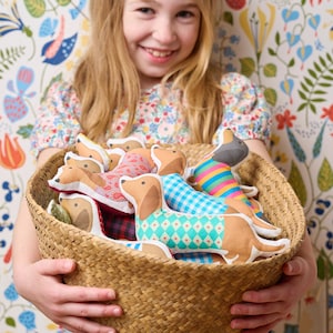 May include: A young girl holds a wicker basket filled with colorful fabric dachshund dog toys. The toys are made of different fabrics and patterns, including plaid, polka dots, and stripes.
