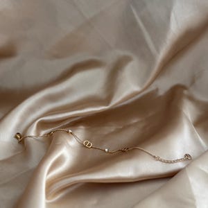 May include: A delicate gold-toned bracelet with small, sparkling accents and the letters "CD" in the center. The bracelet rests on a backdrop of smooth, champagne-colored satin fabric, creating a luxurious aesthetic.