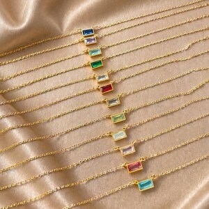 May include: Gold-toned necklaces with rectangular, colorful gemstones. Each necklace features a delicate chain and a single, vibrant stone in shades of blue, purple, green, clear, red, light green, pink, and turquoise. The necklaces are displayed on a soft, golden fabric.