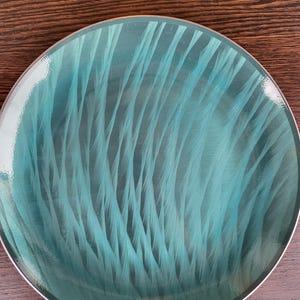 May include: A round teal plate with a silver rim. The plate's surface features a pattern of vertical, feathery lines in varying shades of teal, creating a textured appearance. The plate is set against a dark wood surface.