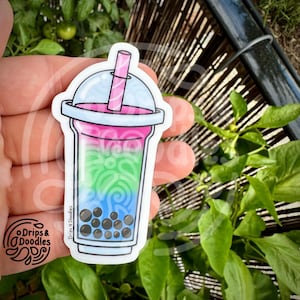 May include: A vibrant sticker depicting a bubble tea. The drink is layered with pink, green, and blue colours, topped with a clear lid and a pink straw. The bottom of the cup has dark brown boba pearls. The sticker has the text "Drips & Doodles".