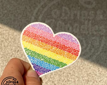 Può includere: Un adesivo a forma di cuore arcobaleno con un motivo a scarabocchi. L'adesivo è composto da diversi colori dell'arcobaleno, tra cui rosso, arancione, giallo, verde, blu, indaco e viola.