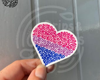 Può includere: Un adesivo bianco a forma di cuore con un design a spirale rosa, viola e blu. L'adesivo è tenuto da una mano.