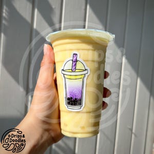 May include: A plastic cup filled with a yellow drink, sealed with a clear lid. A sticker of a boba tea drink with purple and yellow gradients and black boba pearls is on the cup. The sticker has a purple straw.