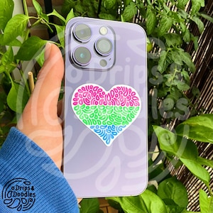 May include: A lilac smartphone with a heart-shaped sticker. The sticker features pink, green, and blue sections, each with a doodle design. The phone has a triple-lens camera.
