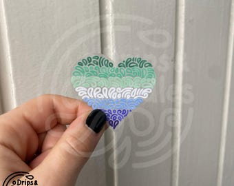 Puede incluir: Una pegatina en forma de corazón verde, azul y morado con un patrón de espiral. La pegatina está sobre una superficie de madera blanca.