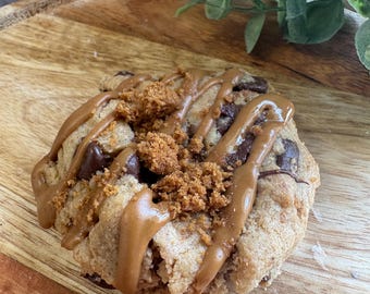 May include: A large cookie with chocolate chips, caramel drizzle, and cookie crumble topping. The cookie sits on a wooden serving platter. The cookie is freshly baked.