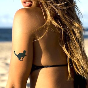 Peut inclure: Deux tatouages ​​temporaires noirs en forme de silhouette de chat sur le bras et le bas du dos. Les tatouages ​​sont sur la peau d'une personne, avec un fond de plage. La personne porte un bikini noir.
