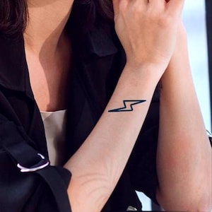 Peut inclure: Tatouage d'éclair noir sur l'avant-bras. Le tatouage est un motif simple. La personne porte une veste noire et a du vernis à ongles rouge. L'image est un gros plan.