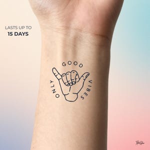 Peut inclure: Un tatouage temporaire représentant un geste de la main avec les mots "Good Vibes Only" dans un design circulaire. Le texte "Lasts up to 15 days" est également inclus. Le tatouage est en noir et blanc.