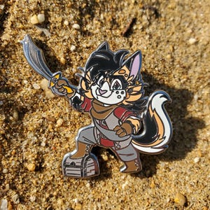 May include: A colorful enamel pin featuring a cartoon character with a sword. The character has black and orange fur, a white face, and is dressed in pirate-themed clothing. The pin is set against a sandy background.