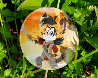 May include: A round sticker featuring a cartoon cat character in a pirate outfit, holding a sword. The cat has orange and black fur, wearing a red jacket and gray pants. The background is orange and yellow.