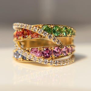 May include: A gold ring with a multi-colored stone design. The ring features a band of small, clear stones with a row of pink, red, and green stones set in the band.
