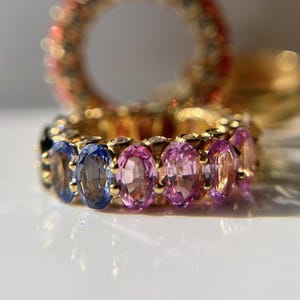 May include: A gold ring with a row of oval-shaped gemstones. The gemstones are a gradient of blue, purple, and pink.