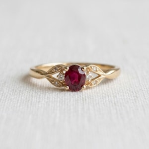 May include: A gold ring featuring a central oval-cut deep red ruby gemstone flanked by two small round-cut white diamonds on each side, set within a polished gold band with a leaf-like design extending from the centre setting along the shank.