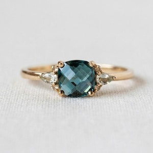May include: A gold ring featuring a central cushion-cut teal gemstone flanked by two smaller pear-cut clear stones. The setting includes small round accent stones surrounding the central gem, all mounted on a polished gold band.