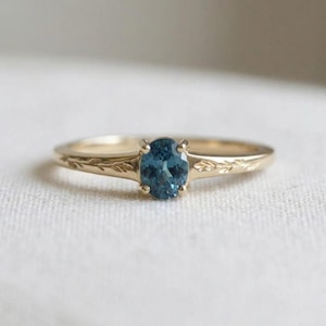 Puede incluir: Un anillo de oro con una piedra preciosa azul de corte ovalado en un engaste de cuatro garras. La banda está hecha de oro amarillo y presenta un patrón de hojas grabado a ambos lados de la piedra central, extendiéndose a lo largo del anillo.