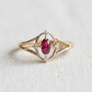 Può includere: Un anello in oro con una gemma centrale di colore rosa intenso dal taglio ovale, inserita in una montatura aperta e allungata. La montatura è impreziosita da quattro piccole pietre chiare dal taglio rotondo sui lati e alle estremità.