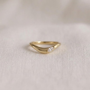 Peut inclure: Une bague en or avec un anneau lisse et incurvé, ornée d'une unique pierre précieuse transparente taille marquise placée de manière décentrée. Le métal présente une finition polie et réfléchissante, formant une forme ondulée continue.