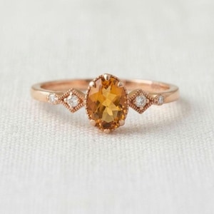 Könnte beinhalten: Ein Ring aus Roségold mit einem zentralen, oval geschliffenen Citrin in einer Krappenfassung. Die Ringschiene ist auf beiden Seiten mit zwei kleinen, runden, klaren Steinen verziert, die in einer rautenförmigen Metallfassung sitzen.