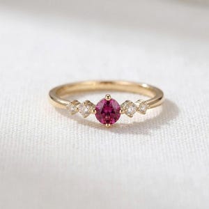 Peut inclure: Une bague en or avec une pierre précieuse rose vif de taille ronde au centre, flanquée de deux pierres blanches transparentes plus petites de chaque côté. L'anneau est en or jaune poli et les cinq pierres sont alignées horizontalement.