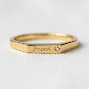 May include: A gold band ring featuring a flat, rectangular front surface engraved with the name Jacob in a cursive script, accompanied by a small, clear, round-cut gemstone set within a star-shaped metal inlay on the right side of the name.
