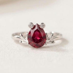 May include: A silver ring featuring a central pear-shaped deep red gemstone. The setting includes two small round clear stones positioned above the pear-shaped gem, with silver prongs extending outward from the base of the central stone.
