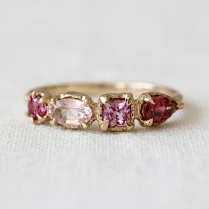 Peut inclure: Une bague en or ornée de quatre pierres précieuses serties en ligne. Les pierres comprennent une gemme ronde rose foncé, une ovale rose clair, une carrée rose vif et une en forme de poire rose foncé, sur un anneau en or poli.