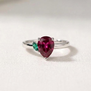 Peut inclure: Une bague argentée avec une pierre précieuse taille poire d'un rouge rosé profond comme point central. À gauche de la pierre principale, une petite pierre verte taille marquise est sertie, rappelant une feuille. L'anneau est lisse et poli.