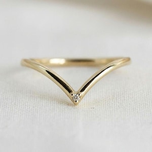 Könnte beinhalten: Ein polierter, V-förmiger Goldring mit einem einzelnen kleinen, runden, klaren Edelstein, der in der Mitte des Chevron-Designs gefasst ist. Das Band hat ein glattes, abgerundetes Profil und eine helle, metallische Oberfläche für ein schlichtes Design.