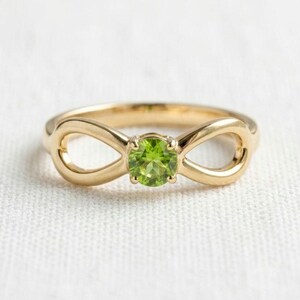May include: A gold ring featuring an infinity symbol design with a round, faceted, lime-green peridot gemstone set in the centre. The band is smooth and polished, showcasing a continuous loop structure that holds the vibrant green stone securely.