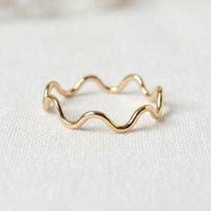 May include: A thin gold ring featuring a continuous wavy or serpentine design. The band has a smooth, polished metallic finish and maintains a consistent thickness throughout its circular shape, creating a delicate and simple jewelry piece.