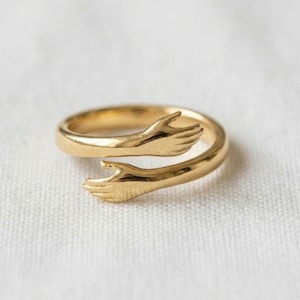 May include: A gold-toned ring featuring a wrap-around design where the two ends are shaped like hands reaching toward each other. The band has a smooth, polished finish and an open structure that allows for adjustment to fit different finger sizes.