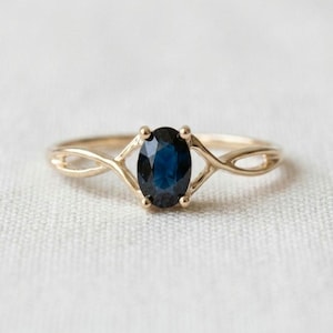Peut inclure: Une bague en or avec un saphir bleu foncé taille ovale au centre, maintenu par quatre griffes. L'anneau est en or jaune poli et se divise en deux brins délicats et entrelacés de chaque côté de la pierre, formant un design croisé.