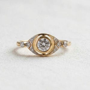 Puede incluir: Un anillo de oro con un diamante central de talla brillante engastado en un bisel circular. La banda está decorada con pequeños diamantes incrustados en formas metálicas geométricas a ambos lados, creando un diseño alargado en forma de ojo.