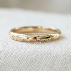 Könnte beinhalten: Ein goldener Bandring mit polierter Oberfläche, gravierten Sternmotiven und kleinen, runden, klaren eingesetzten Steinen. Das Design umfasst eine zentrale sternförmige Fassung mit einem größeren Stein, umgeben von kleineren Sternen und Akzentsteinen.