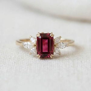 以下が含まれることがあります： 中央にエメラルドカットの深い赤色の宝石をあしらったゴールドリング。中央の石の両サイドには、マーキスカットの透明な白い石が3つずつ扇状に配置されており、磨き上げられたゴールドのバンドにセットされています。
