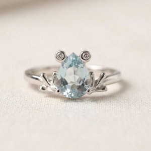 Könnte beinhalten: Ein silberfarbener Ring mit einem zentralen, birnenförmigen hellblauen Edelstein. Die Fassung enthält zwei kleine runde Akzentsteine an der Oberseite und dekorative Metallkrallen an der Basis, die an ein krabbenartiges Design erinnern.