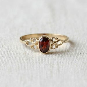 Könnte beinhalten: Ein goldener Ring mit einem zentralen, oval geschliffenen, tiefroten Granat-Edelstein. Die Ringschiene ist auf beiden Seiten des Steins mit filigranen, durchbrochenen Schnörkel-Metalldetails verziert, die dem Schmuckstück ein zartes Aussehen verleihen.