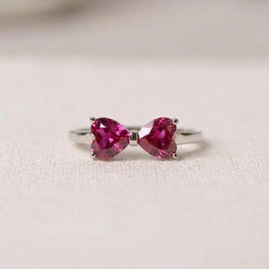 May include: A silver-toned ring featuring two vibrant, heart-shaped, deep pink gemstones arranged to form a bow shape. The band is smooth and metallic, connecting to the center where the two faceted stones meet at their points to create a bow design.