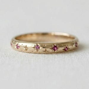 Puede incluir: Un anillo de banda de oro con una serie de patrones de estrellas grabados. Varias de estas estrellas están engastadas con pequeñas piedras preciosas redondas de color rosa vibrante. La superficie del metal es pulida y lisa, con un diseño celestial.