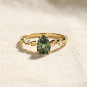 May include: A gold ring featuring a pear-shaped green gemstone at the centre, flanked by two small clear stones. The band is designed with a delicate vine and leaf motif that extends from the central setting along both sides of the ring.