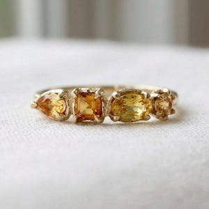 Puede incluir: Un anillo de oro con cuatro piedras preciosas amarillas en fila. Las piedras están dispuestas horizontalmente e incluyen una talla pera, una cuadrada, una ovalada y una pequeña redonda, todas aseguradas con garras de metal en una banda fina.