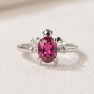 Op de afbeelding: Een zilverkleurige ring met een centrale, ovaal geslepen, felroze edelsteen. De zetting heeft de vorm van een schildpad, waarbij de edelsteen het schild vormt en kleine metalen uitsteeksels de kop en de vier poten van het dier weergeven.
