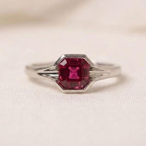 May include: A silver-toned ring featuring a central, square-cut, deep pinkish-red gemstone set in a bezel setting. The band has a tapered design that widens as it reaches the stone, creating a sleek, polished look with clean, metallic lines.