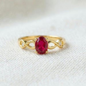 Puede incluir: Un anillo de oro con un rubí rojo intenso de talla ovalada en el centro, sujeto por un engaste de cuatro garras. La banda presenta un diseño de infinito a ambos lados, con una pequeña piedra de acento redonda y clara en cada bucle.
