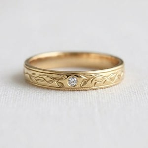 Könnte beinhalten: Ein goldener Ring mit einem eingravierten Ranken- und Blattmuster, das den gesamten Außenbereich umschließt. Ein einzelner kleiner, runder, klarer Edelstein ist in die Mitte des Rings eingelassen und direkt in die Metalloberfläche integriert.