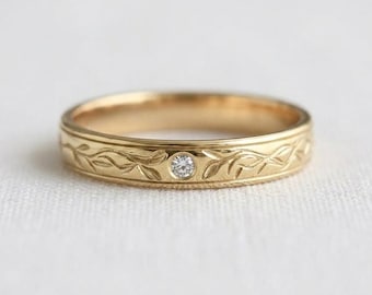 May include: A gold band ring featuring an engraved vine and leaf pattern encircling the exterior. A single small, round, clear gemstone is set into the center of the band, integrated directly into the metal surface of the ring.